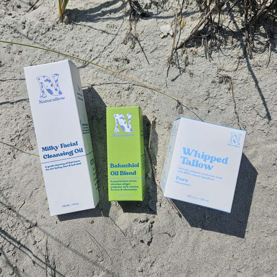 Three skincare product boxes from Naturallow arranged on sandy ground outdoors: Milky Facial Cleansing Oil, Bakuchiol Oil Blend, and Whipped Tallow. The boxes are white and light green, with blue text and logos, surrounded by sand and dry grass.