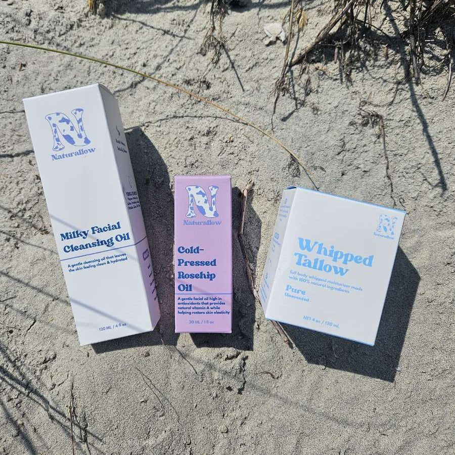 Three skincare product boxes from Naturallow laid out on sandy ground outdoors: a white Milky Facial Cleansing Oil box, a pink Cold-Pressed Rosehip Oil box, and a white Whipped Tallow box.