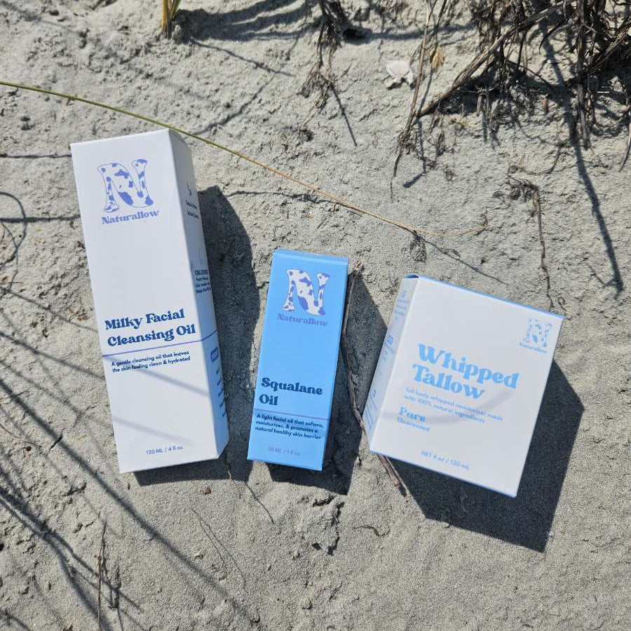 Three skincare product boxes from Naturallow are arranged on sandy dune grass. The products include Milky Facial Cleansing Oil in a tall white box, Squalane Oil in a blue box, and Whipped Tallow in a white box.
