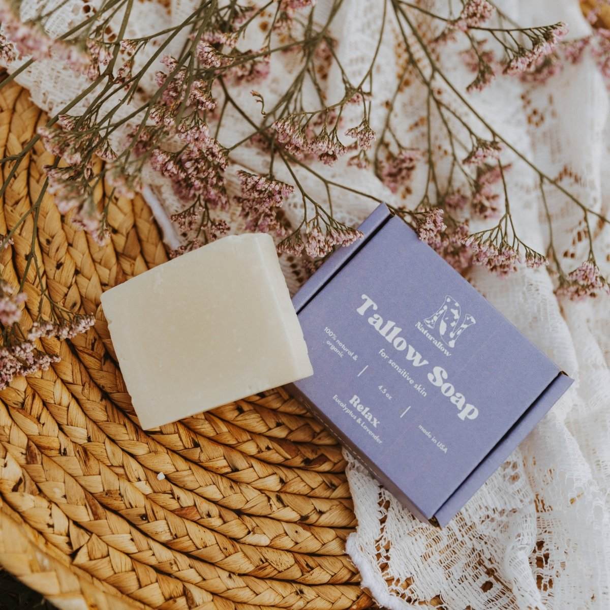 Naturallow Relax Tallow Soap bar propped up next to the box on a white lace cloth on top of a woven wooden basket with flowers