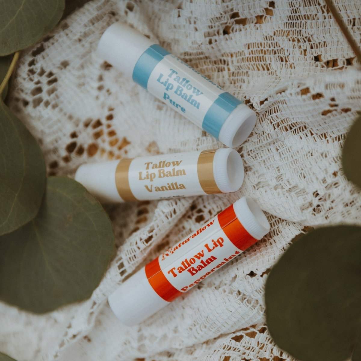 3 of Naturallow's Vanilla, Peppermint, and Pure Unscented Tallow Lip Balms Spread Out On A White Lace Surface