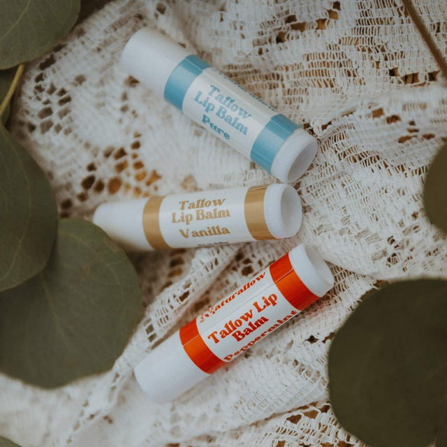 3 of Naturallow's Vanilla, Peppermint, and Pure Unscented Tallow Lip Balms Spread Out On A White Lace Surface