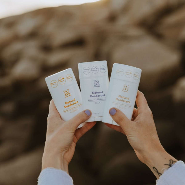 A woman holding up 3 of Naturallow's Natural Deodorants, Citrus, Relax, and Vanilla, next to each other against the horizon
