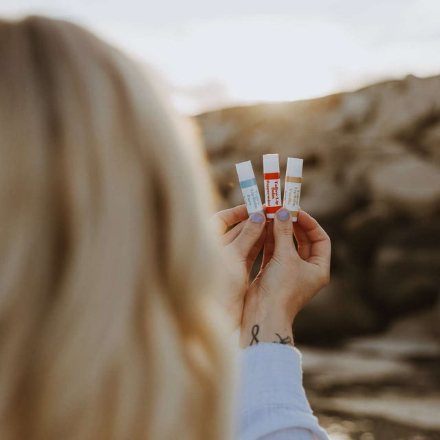 The back of a womans head as she holds up 3 of Naturallow's Vanilla, Peppermint, and Pure Unscented Tallow Lip Balms against the horizon