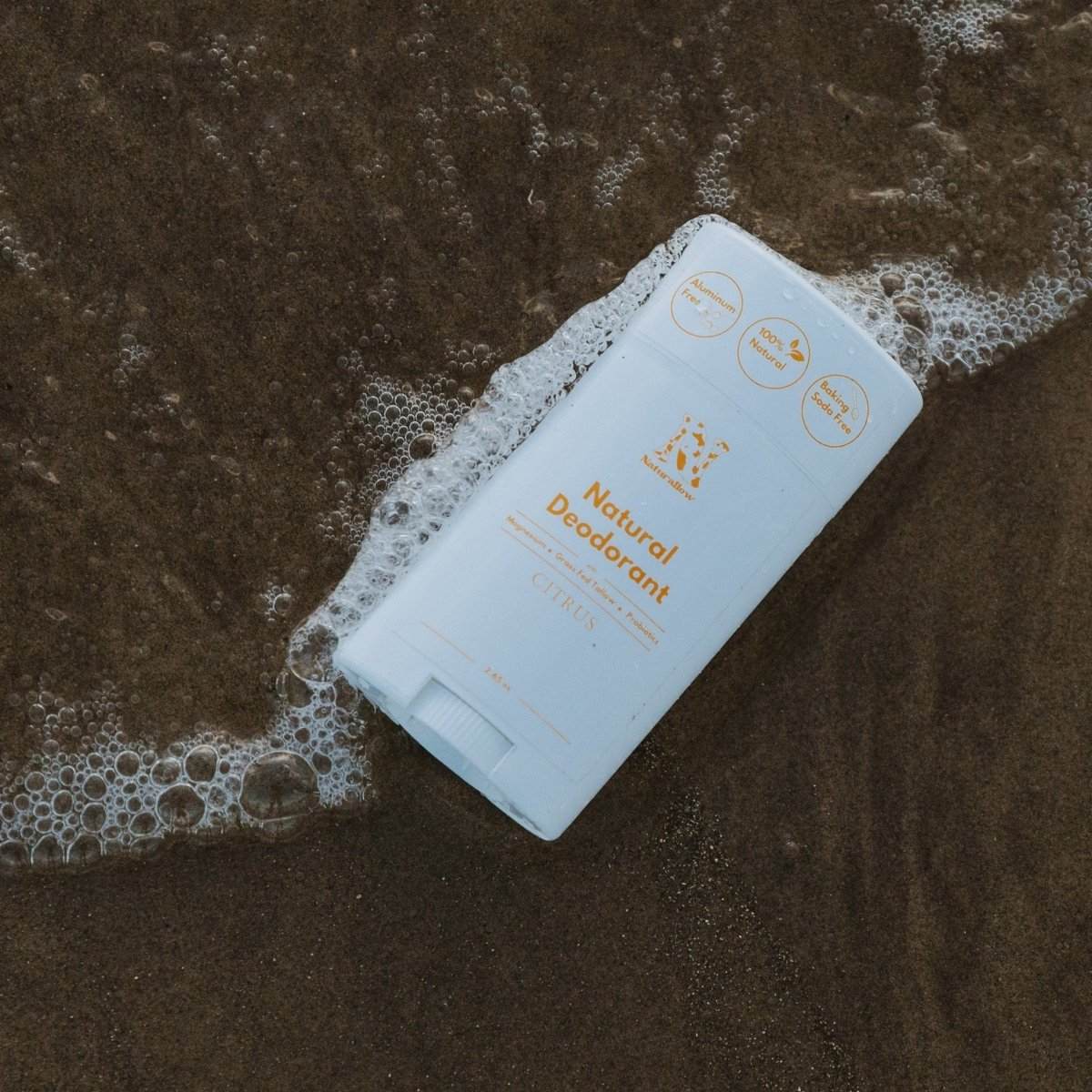 Naturallow's Citrus Natural Deodorant lying on the sand in the shallow beach surf