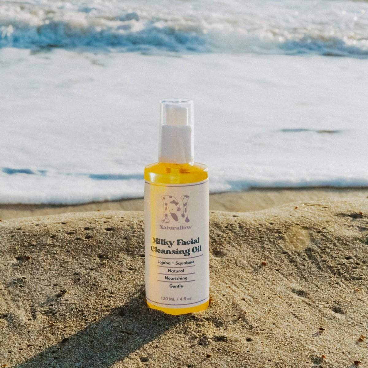 A close-up of Naturallow's Milky Facial Cleansing Oil bottle sitting by itself on the beach with the ocean in the background