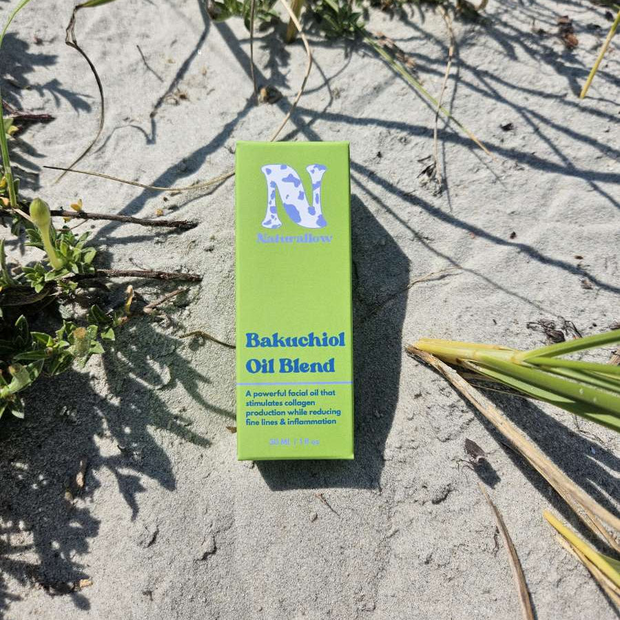A small green box of Naturallow Bakuchiol Oil Blend lying on sandy ground with some grass and shadows around it.