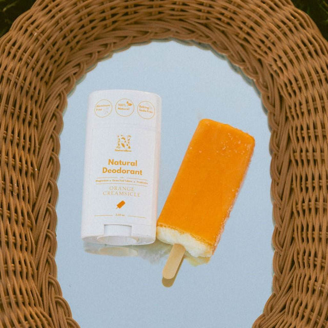 Naturallow's Limited Edition Orange Creamsicle deodorant lying next to a orange popsicle on a mirrored surface