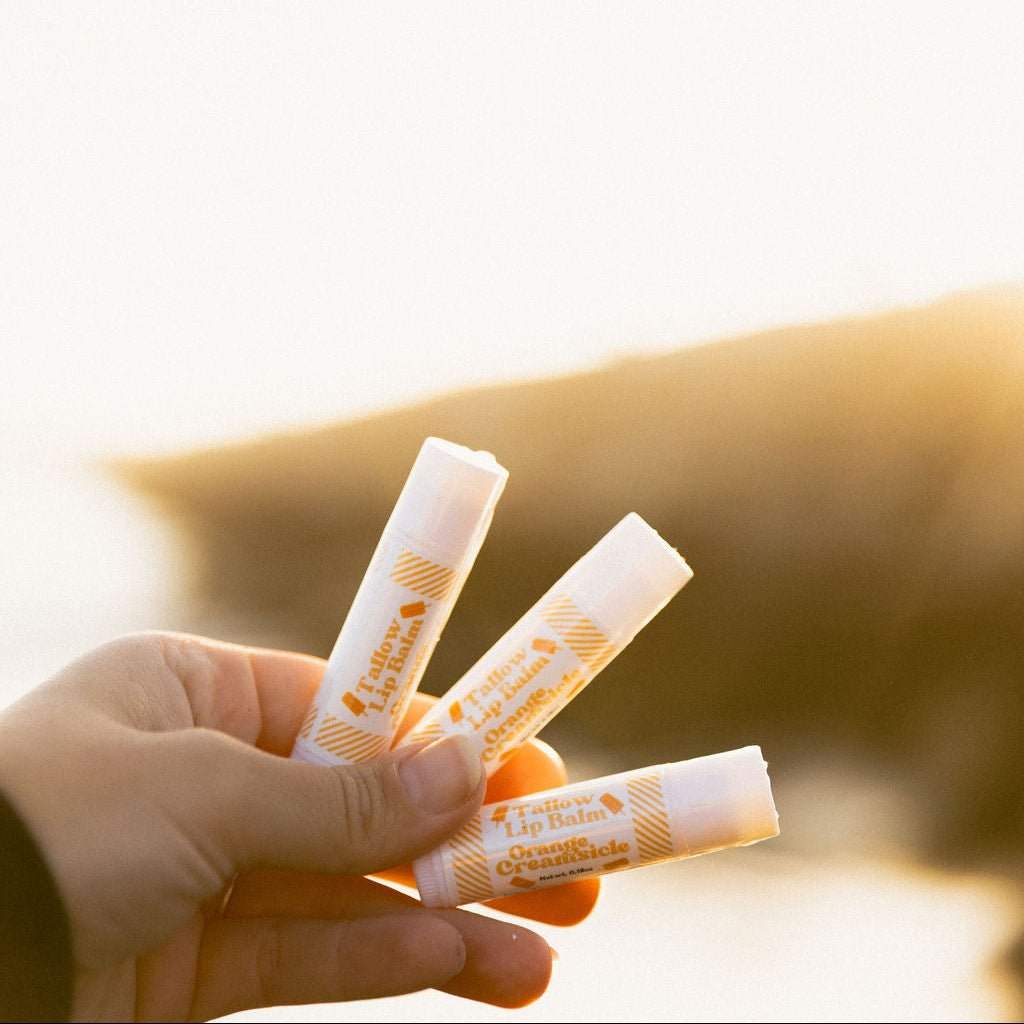 A hand holding three of Naturallow's Limited Edition Orange Creamsicle Tallow Lip Balms against the horizon