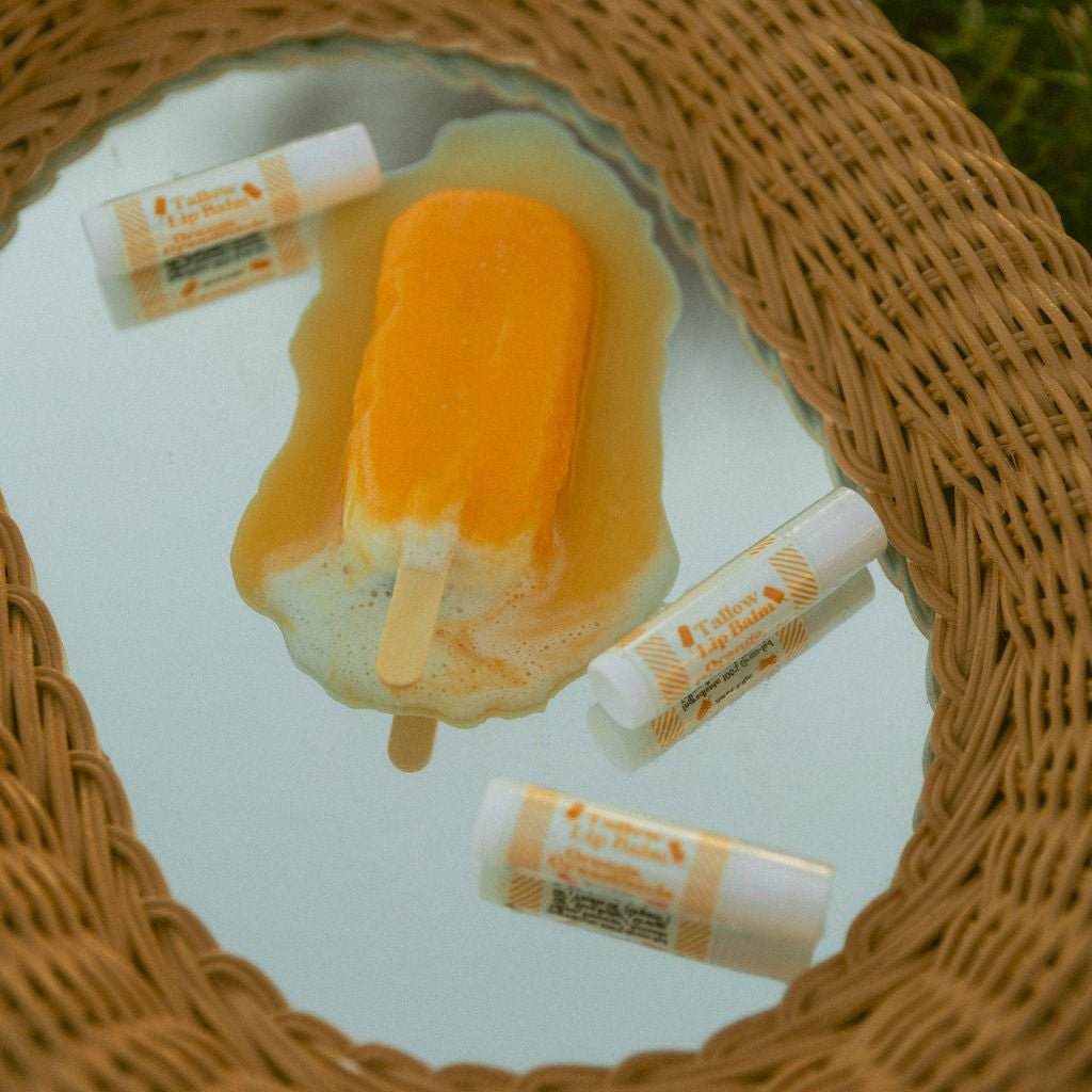 Three of Naturallow's Limited Edition Orange Creamsicle Tallow Lip Balms on laying around a melting orange popsicle on top of a mirrored surface