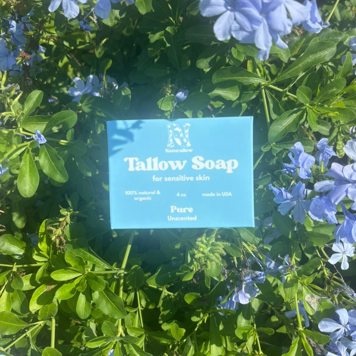 A light blue rectangular Naturallow soap bar labeled 'Tallow Soap for sensitive skin' placed among green leaves and small light blue flowers outdoors.