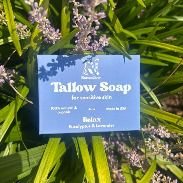 A purple rectangular soap box labeled 'Tallow Soap' by Naturallow, with 'Relax' as the scent name, nestled among green leaves and small purple flowers outdoors.