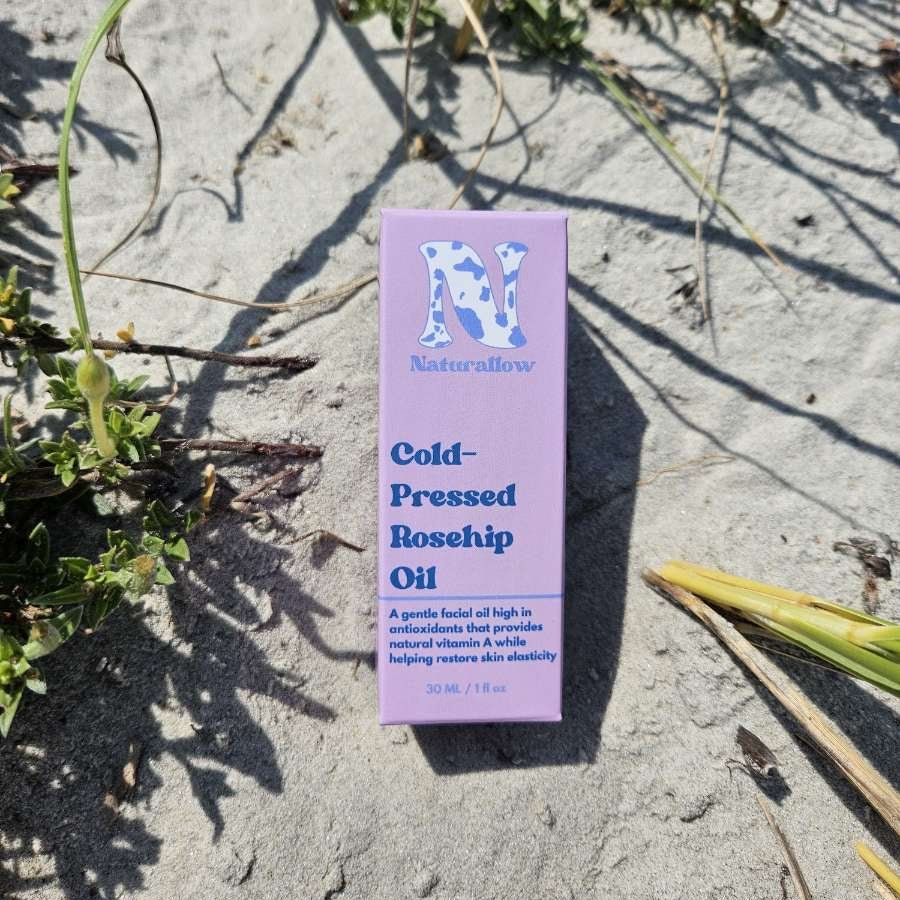 A pink box of Naturallow Cold-Pressed Rosehip Oil placed on sandy ground outdoors, surrounded by small green plants and casting a shadow.