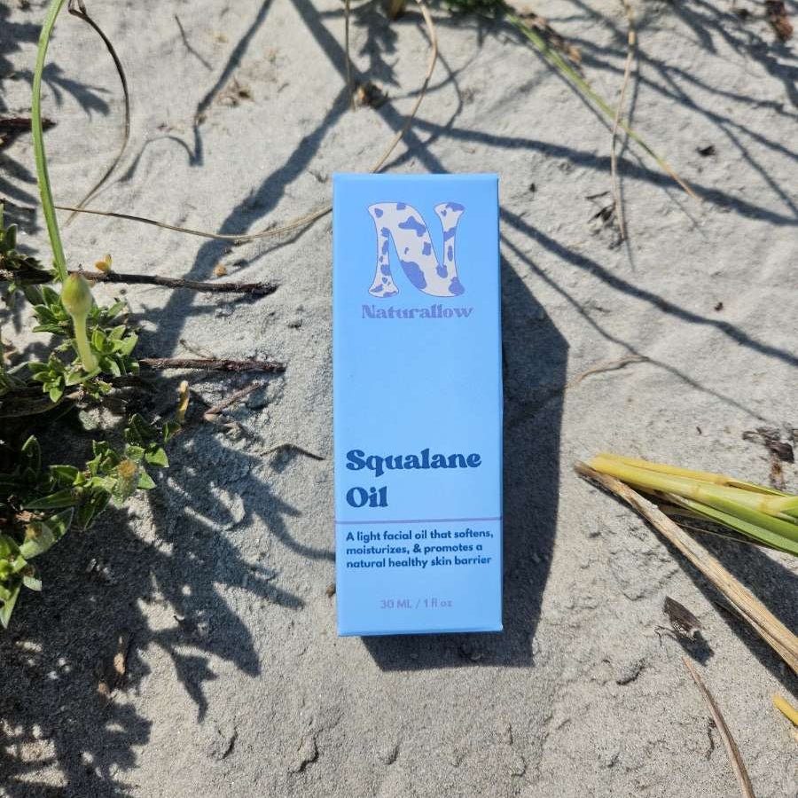 A light blue rectangular box of Naturallow Squalane Oil for dry, sensitive, and sun-damaged skin, placed on sandy ground with some green beach grass.